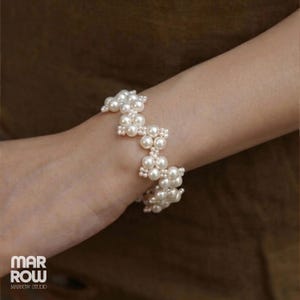Peut inclure: Un bracelet composé de perles artificielles blanches sphériques disposées en un motif répétitif en zigzag ou en chevron. Les perles sont reliées pour former une bande géométrique structurée autour du poignet. Le texte MAR ROW MARROW STUDIO est visible.