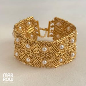 Peut inclure: Un bracelet doré en perles avec un motif répété de petites perles blanches. Le bracelet est composé de petites perles dorées tissées ensemble pour créer une large bande. Le fermoir est doré et comporte une petite chaîne.