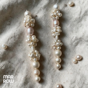 May include: A pair of long drop earrings featuring clusters of small white freshwater pearls interspersed with larger, smooth, round, off-white and pale pink pearls. The earrings are arranged in a vertical, multi-layered design.