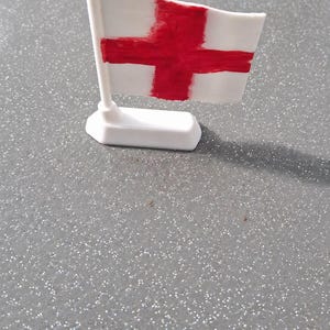 May include: A miniature flag of England, featuring a white background with a red St George's cross. The flag is mounted on a white pole and base. The flag appears to be made of a textured material, with the cross painted on.
