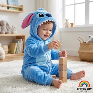 Könnte beinhalten: Ein Baby in einem blauen Plüsch-Overall mit Stitch-Design, komplett mit einer Kapuze mit großen Augen und einem grinsenden Mund. Das Kind spielt mit Holzblöcken auf einem weißen Teppich. Das Little Joy Studio-Logo befindet sich unten rechts.