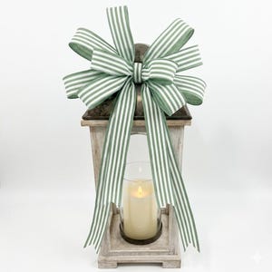 May include: A distressed wooden lantern with a battery-operated candle inside, adorned with a large green and white striped bow. The lantern features a weathered finish, and the candle is within a clear glass cylinder.