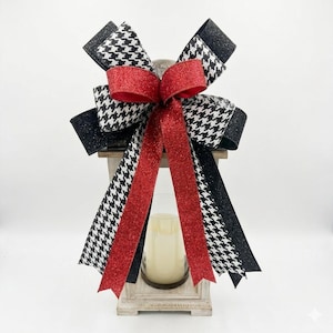 May include: A decorative lantern topped with a large bow. The bow combines black and white houndstooth and solid black ribbon, with a central red glitter ribbon. A candle is visible inside the lantern.
