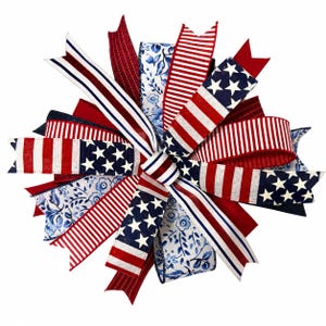 May include: A decorative bow made of multiple wired ribbons featuring red, white, and blue color schemes. The patterns include stars and stripes, solid red, and a blue and white floral print. The ribbons are arranged in a circular starburst shape.