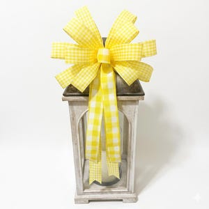 May include: A white wooden lantern adorned with a yellow and white gingham bow. The lantern features a metal top and a candle inside. The bow is large and decorative, with long, flowing ribbons. The lantern is a decorative item.