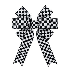 May include: A decorative bow crafted from fabric featuring a black and white chequered pattern. The bow consists of multiple loops and two long, downward-hanging tails with notched ends, all displaying the consistent square grid design.
