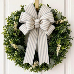 May include: A circular green wreath with a cream-coloured bow featuring blue vertical stripes. The wreath is composed of green foliage and small white flowers. It is suspended on a white door.