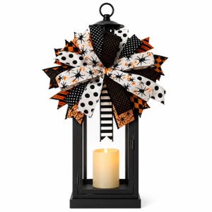 Könnte beinhalten: Schwarze Metalllaterne mit einer dekorativen Schleife. Die Schleife hat Bänder in Schwarz, Weiß und Orange mit Mustern wie Punkten, Spinnen und Streifen. Eine cremefarbene Kerze befindet sich in der Laterne, ideal für Halloween. Höhe ca. 30 cm.