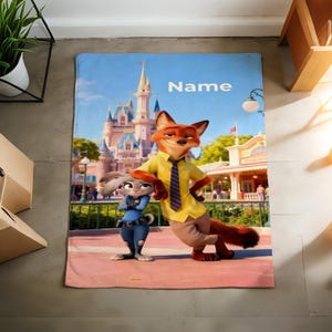 May include: A rectangular blanket featuring the characters Nick Wilde and Judy Hopps from Zootopia, with a castle backdrop and the word "Name" in white. The blanket has a pink border and a blue sky background.