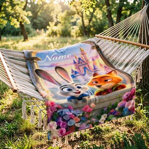 May include: A personalised blanket featuring cartoon characters Judy Hopps and Nick Wilde from Zootopia, with a castle backdrop and floral border. The blanket is draped over a hammock in a sunny outdoor setting. The word "Name" is visible.