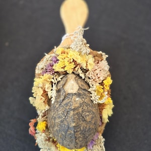 May include: A wooden scoop decorated with an arrangement of dried flowers in shades of yellow, purple, and cream, surrounding a dark, textured, oval-shaped turtle shell positioned in the center of the scoop's bowl.
