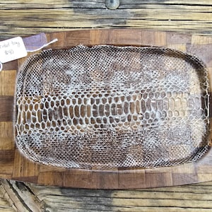 May include: A rectangular wooden tray with a checkered pattern featuring a central inlay of textured brown and white snakeskin. A small white paper tag is attached to the corner with the handwritten text "trinket tray $40".