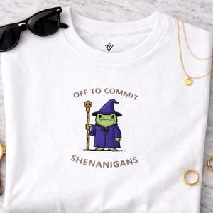 Może przedstawiać: Biała koszulka z napisem "OFF TO COMMIT SHENANIGANS" i kreskówkową żabą ubraną w strój czarodzieja. Żaba jest zielona, ma fioletową szatę i kapelusz, trzymając laskę. Widoczne są okulary przeciwsłoneczne, zegarek i złota biżuteria.