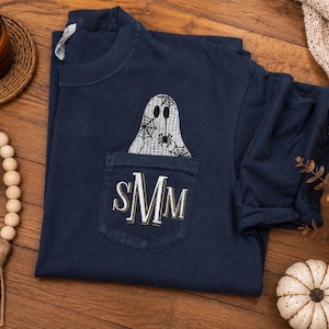 May include: A navy blue short-sleeve t-shirt with a chest pocket. The pocket features a white embroidered monogram with the letters SMM. Above the pocket, a white ghost graphic with a spider web pattern and a small spider is embroidered.