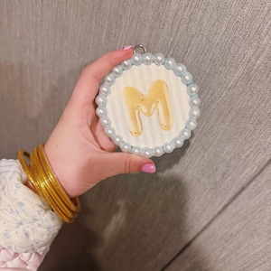 May include: A round, decorative ornament featuring the letter "M" in a gold, bubble-like font. The ornament has a light blue and white striped background, surrounded by a border of small, white pearls. A gold bangle bracelet is visible.