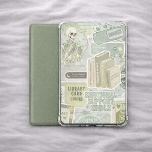 Könnte beinhalten: Eine grüne Kindle-Hülle mit einer transparenten Hülle, die eine Collage von Aufklebern zeigt. Die Aufkleber zeigen ein Skelett, Bücher und Text mit der Aufschrift "Emotional Support Kindle" und "Library Card". Die Hülle befindet sich neben einer grünen Abdeckung.