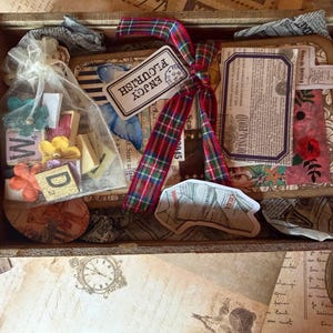 May include: A wooden box containing craft materials. It includes a small journal, a bag of wooden letter blocks, and various decorative items. The journal is tied with a tartan ribbon and a tag that reads "ENJOY FLOURISH".