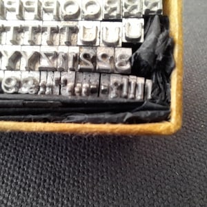 May include: A vintage metal type set with individual letters and numbers arranged in rows within a wooden box. The metal type is silver-colored, and the box has a gold-colored trim. The set includes various fonts and sizes for printing.