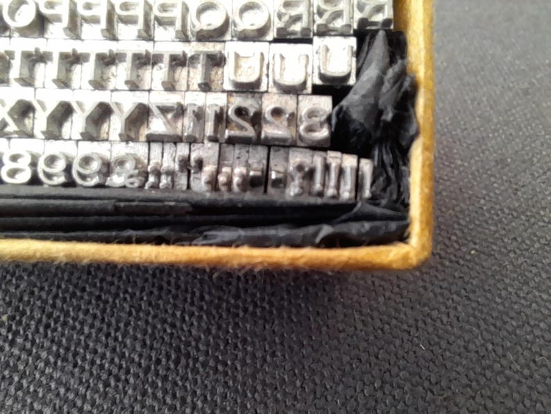 May include: A vintage metal type set with individual letters and numbers arranged in rows within a wooden box. The metal type is silver-colored, and the box has a gold-colored trim. The set includes various fonts and sizes for printing.