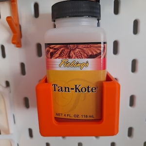 May include: A bright orange plastic holder mounted on a white pegboard, containing a bottle of Fiebing's Tan-Kote leather finish. The bottle has a black cap, a yellow and brown label with the text Tan-Kote, and states a volume of 4 fl. oz.