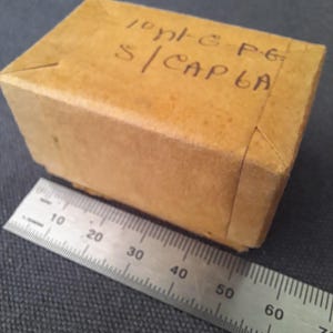 May include: A small, rectangular cardboard box with handwritten text on the top. A metal ruler is placed next to the box, showing measurements in centimeters. The box is a light brown color.