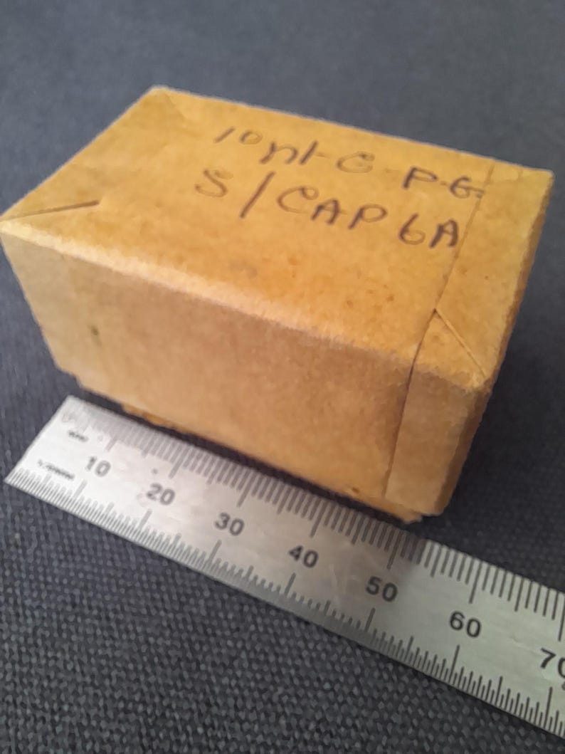 May include: A small, rectangular cardboard box with handwritten text on the top. A metal ruler is placed next to the box, showing measurements in centimeters. The box is a light brown color.