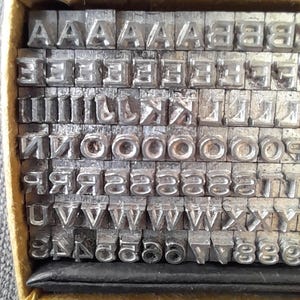 May include: A collection of metal letterpress printing type, including letters and numbers, neatly arranged in a yellow box. The type is made of a silver-colored metal and is used for printing.