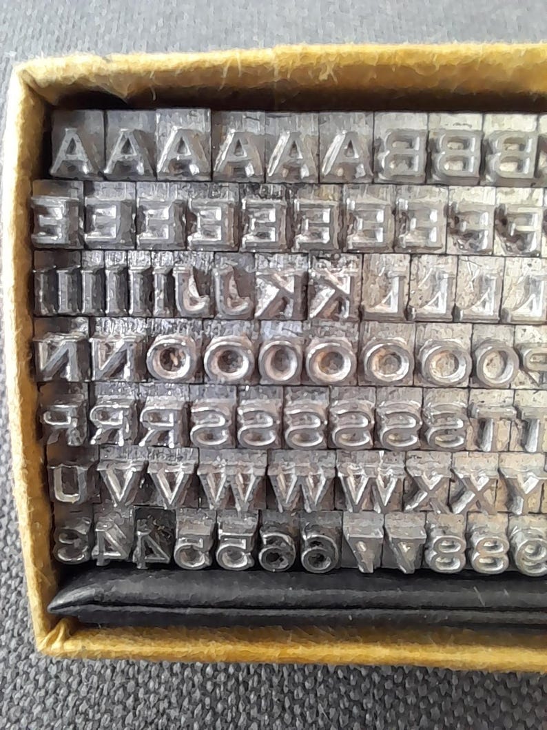 May include: A collection of metal letterpress printing type, including letters and numbers, neatly arranged in a yellow box. The type is made of a silver-colored metal and is used for printing.