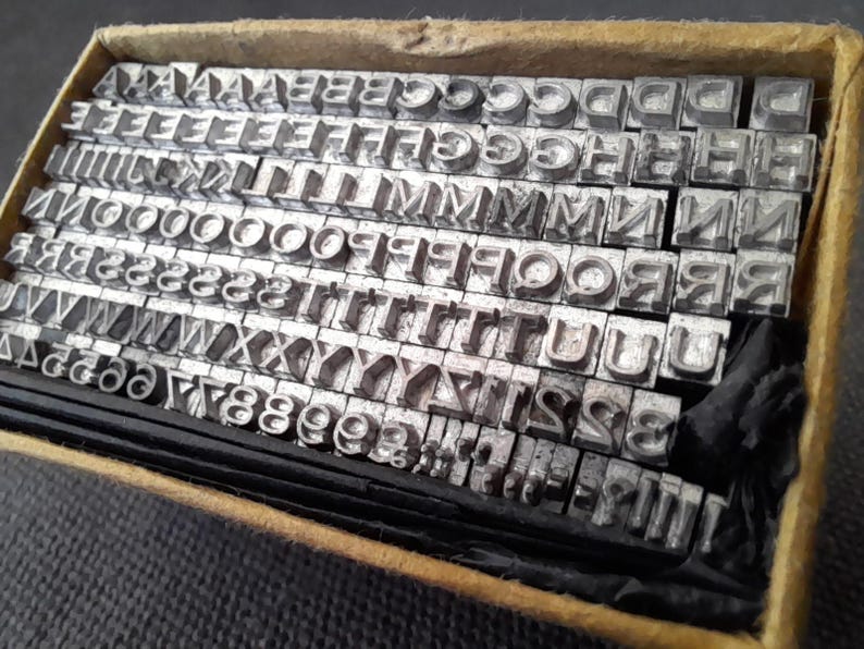 May include: A vintage typeset with metal letter blocks, including letters and numbers, neatly arranged in a cardboard box. The letters are in a serif font, and the box has a worn, aged appearance, suggesting its use in printing or typesetting.