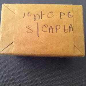 May include: A small, rectangular cardboard box with handwritten text in black ink. The box is a light brown color and appears to be sealed. The text on the box reads "10 nt-G-P-G S | CAP 6A".