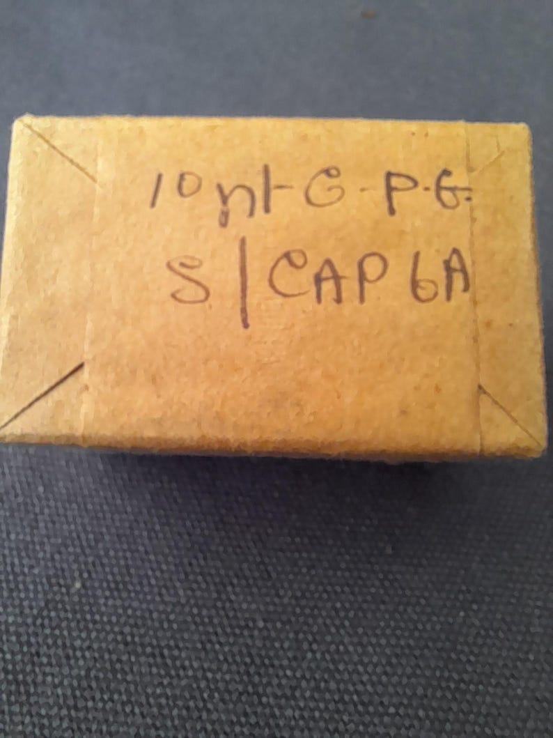 May include: A small, rectangular cardboard box with handwritten text in black ink. The box is a light brown color and appears to be sealed. The text on the box reads "10 nt-G-P-G S | CAP 6A".