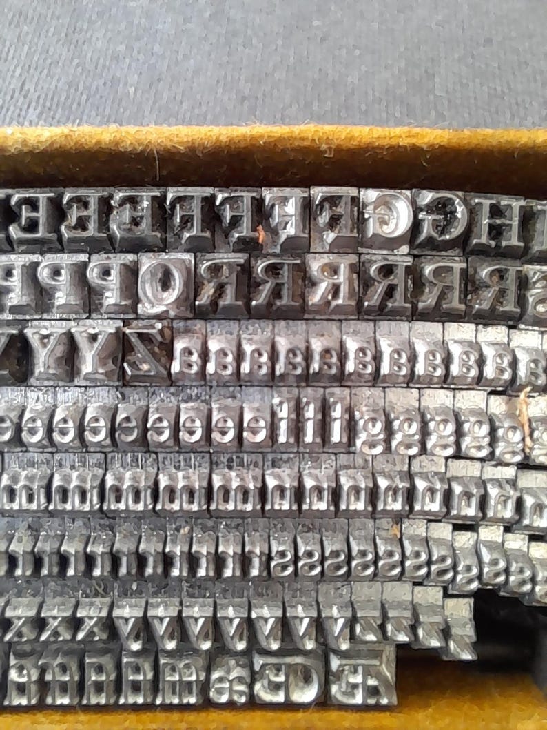 May include: Close-up of a collection of vintage metal letterpress printing type. The letters are arranged in rows within a wooden tray. The metal type is silver-colored and shows signs of wear, indicating its age and use. The tray has a yellow interior.