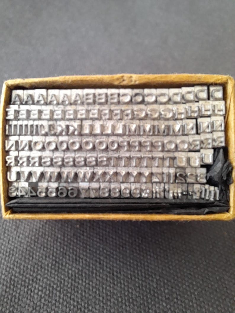 May include: A set of metal letterpress printing type, neatly arranged in a cardboard box. The letters are in a variety of fonts and sizes, perfect for creating custom text. The box is a warm, neutral color, and the type is a silver-gray.