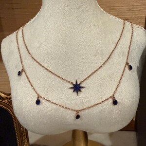 May include: A two-strand necklace displayed on a mannequin. The necklace features a rose gold chain with a dark blue star pendant and several dark blue teardrop-shaped charms. The mannequin is cream-colored and has a lace trim at the neck.