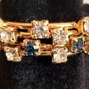 May include: Gold-toned ring with clear and blue gemstones. The ring features a series of connected bands adorned with square and floral-shaped stones. The gemstones are set in a gold metal, creating a vintage or antique aesthetic.