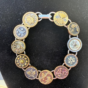 May include: A circular bracelet composed of twelve distinct round buttons linked together by silver-toned metal rings. The buttons feature varied metallic finishes in gold, bronze, and silver tones, displaying embossed patterns including floral motifs, celestial shapes, and geometric designs.