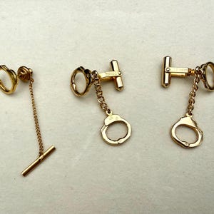 May include: A set of gold-toned jewelry consisting of one tie bar with a chain and a circular charm, and two matching cufflinks. Each piece features a miniature handcuff design connected by a delicate gold chain, showcasing a polished metallic finish.
