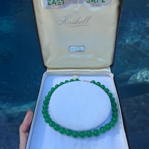 May include: A green beaded necklace presented in a cream-colored display box. The interior lid features the text East Jade by Haskell. The necklace consists of uniform, round, translucent green beads arranged in a circular pattern on a white base.
