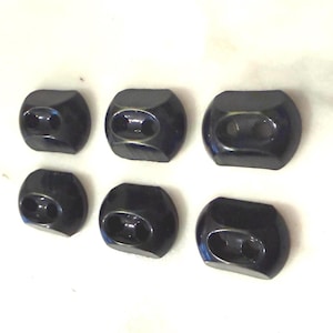 May include: Six black cord locks with a glossy finish are arranged on a white surface. Each lock has a rounded, rectangular shape with two holes. The cord locks are likely used for adjusting drawstrings or cords on clothing or accessories.