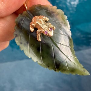 May include: A pendant featuring a carved dark green leaf with a textured gold-tone frog resting on its surface. The frog has two small pink gemstone eyes and a detailed, bumpy skin texture. The leaf has visible carved veins and a smooth finish.