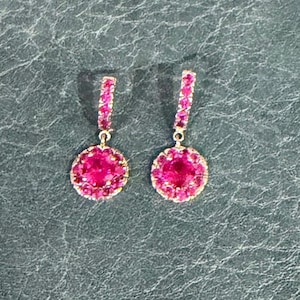 May include: A pair of drop earrings featuring a thin, vertical bar encrusted with small, vibrant pink stones, leading to a circular pendant at the base. The circular pendant is also set with a central pink stone surrounded by a halo of smaller pink stones, all mounted in a metallic setting.