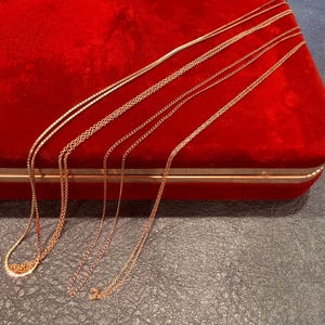 May include: A collection of four delicate rose gold-toned metal chains of varying link styles, including box and cable chain designs, draped across a bright red velvet-lined jewelry box with a gold-toned metal trim.