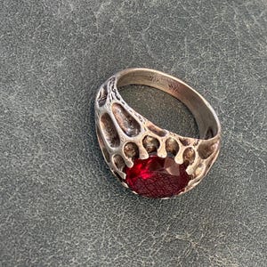 May include: A silver-toned ring featuring an oval-shaped, deep red faceted gemstone set within a textured, organic, web-like metal frame. The band has a weathered, porous appearance with visible hallmarks stamped on the inner surface of the ring.