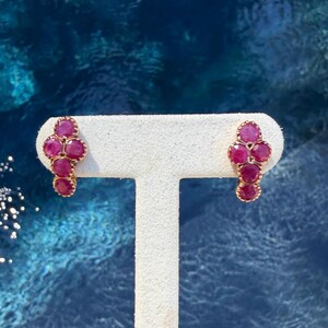 May include: A pair of stud earrings featuring a cluster of four round, deep pinkish-red gemstones set in gold-toned metal. Each earring is arranged in a vertical, cross-like formation, showcasing faceted surfaces that catch the light.