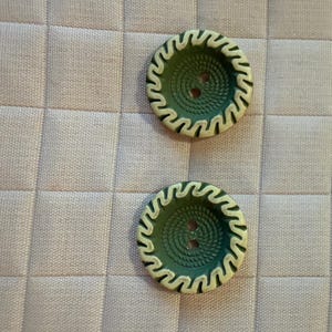 May include: Two round, green buttons with a spiral design and two holes. The buttons have a cream-colored, wavy border. The buttons are on a light pink, quilted fabric.