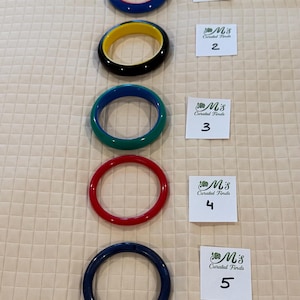 May include: Five colorful bangles in red, blue, green, and black with a yellow interior. Each bangle is approximately 0.5 inches wide. The bangles are arranged vertically on a textured, light-colored surface. Each bangle has a corresponding numbered tag.