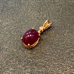 May include: A gold pendant featuring a smooth, oval-shaped deep red cabochon gemstone held in a four-prong setting. A small, clear, round-cut accent stone is mounted on the gold bail at the top of the pendant, which connects to a chain.
