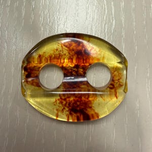 May include: Oval-shaped amber-colored object with two circular holes. The translucent material has reddish-brown inclusions, creating a unique pattern. The object is likely a decorative element or component.
