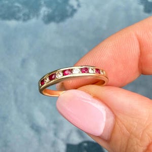 Puede incluir: Un anillo de banda de oro con un engaste de canal que alterna rubíes rojos y diamantes blancos transparentes de corte redondo. Las piedras están dispuestas en una sola fila sobre la parte superior de la banda de metal pulido.