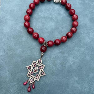 May include: A necklace featuring a strand of faceted deep red beads and a silver-toned geometric pendant. The pendant is embellished with small red stones and clear crystals, with three dangling red teardrop-shaped beads at the bottom.